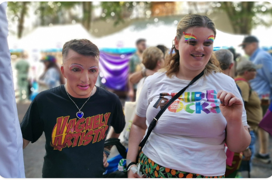 Mark, to the left wears a black tshirt with Vaguely Artistic printed in red capital letters and a yellow shape in the background. They are wearing pink lipstick overlining their lips and colourful pink and white eyeshadow. Mark also has a silver chain with a blue heart-shaped gemstone around their neck. Katie, to the right, is wearing a white tshirt with the words pride rocks on the front, in the colours of the pride flag. They are wearing gemsones on their eyebrows in the colours of the pride flag and the transgender pride flag colours on their lips. Katie is smiling while looking away from the camera.