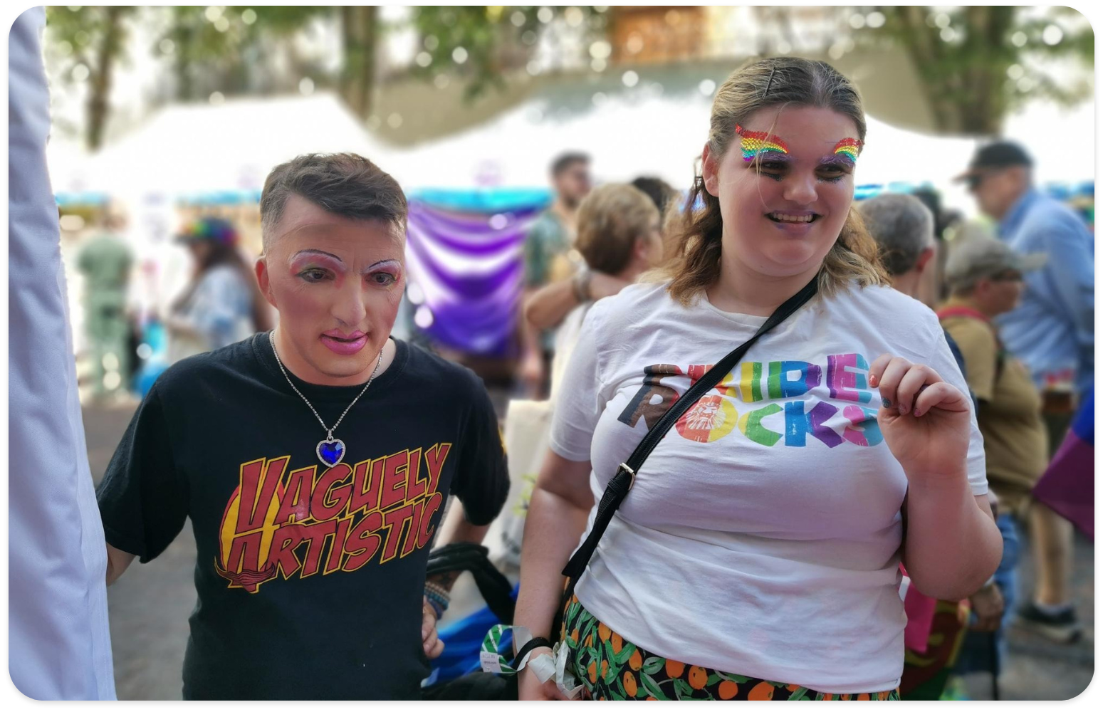 Mark, to the left wears a black tshirt with Vaguely Artistic printed in red capital letters and a yellow shape in the background. They are wearing pink lipstick overlining their lips and colourful pink and white eyeshadow. Mark also has a silver chain with a blue heart-shaped gemstone around their neck. Katie, to the right, is wearing a white tshirt with the words pride rocks on the front, in the colours of the pride flag. They are wearing gemsones on their eyebrows in the colours of the pride flag and the transgender pride flag colours on their lips. Katie is smiling while looking away from the camera.